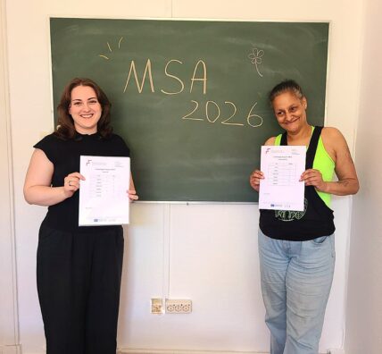 Zwei Frauen stehen vor einer Tafel auf der MSA 2026 steht. Sie halten ein Zeugnis in der Hand und lächeln.