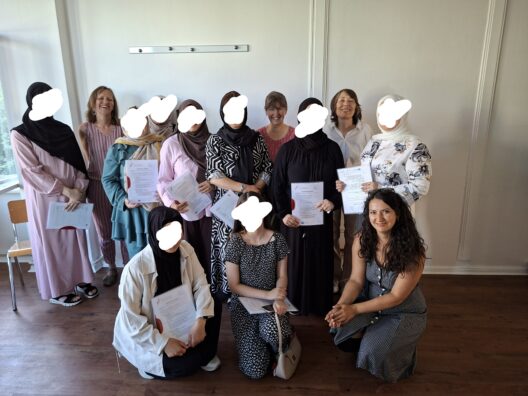 Eine Gruppe Frauen steht zusammen vor einer weißen Wand. Von den Teilnehmerinnen sind die Gesichter anonymisiert, sie halten ihr Zertifikat in der Hand. Vier Mitarbeiterinnen lächeln und stehen mit auf dem Bild.