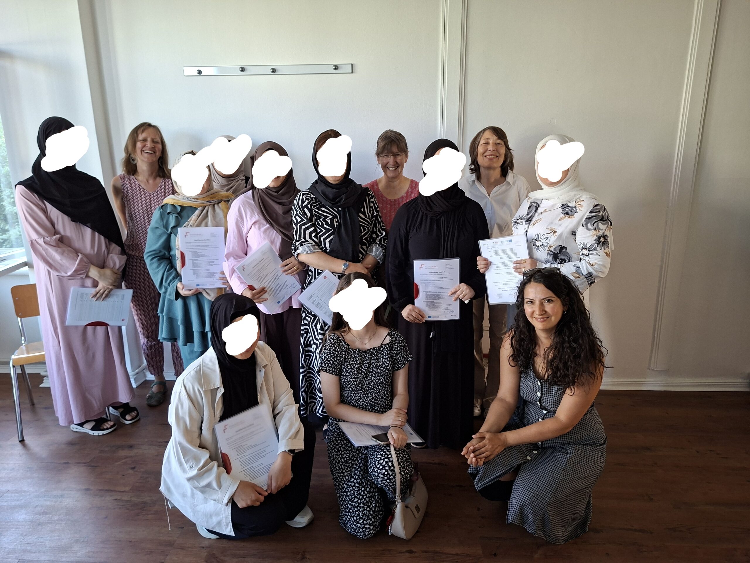 Eine Gruppe Frauen steht zusammen vor einer weißen Wand. Von den Teilnehmerinnen sind die Gesichter anonymisiert, sie halten ihr Zertifikat in der Hand. Vier Mitarbeiterinnen lächeln und stehen mit auf dem Bild.